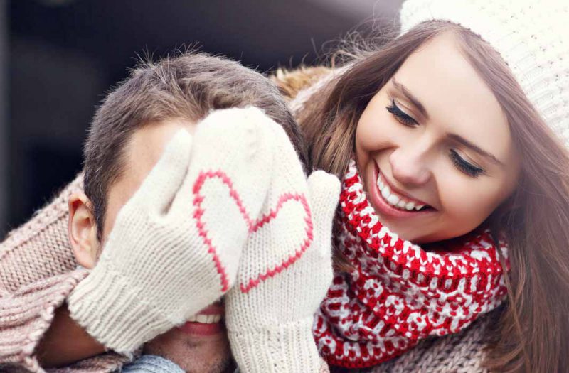 A woman hiding a man's face with wollen gloves with a heart pattern on the front on valentines day